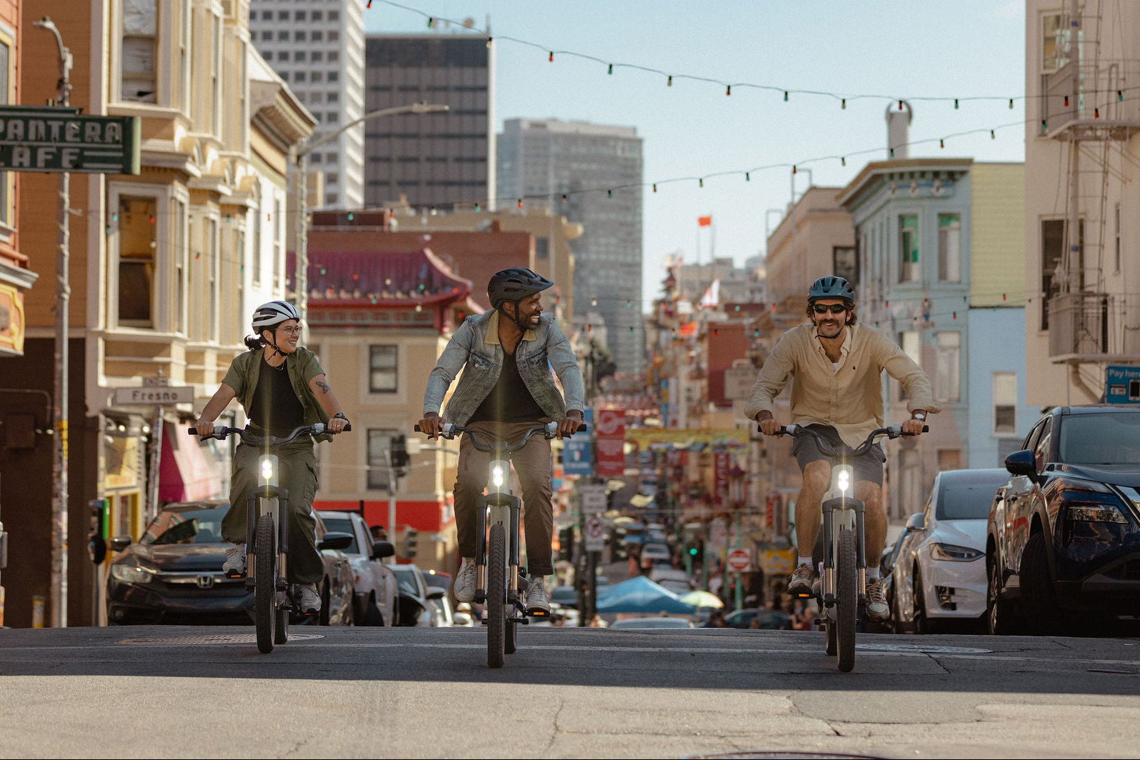 Three people riding bicycles on a city street with tall buildings and cars in the background.
